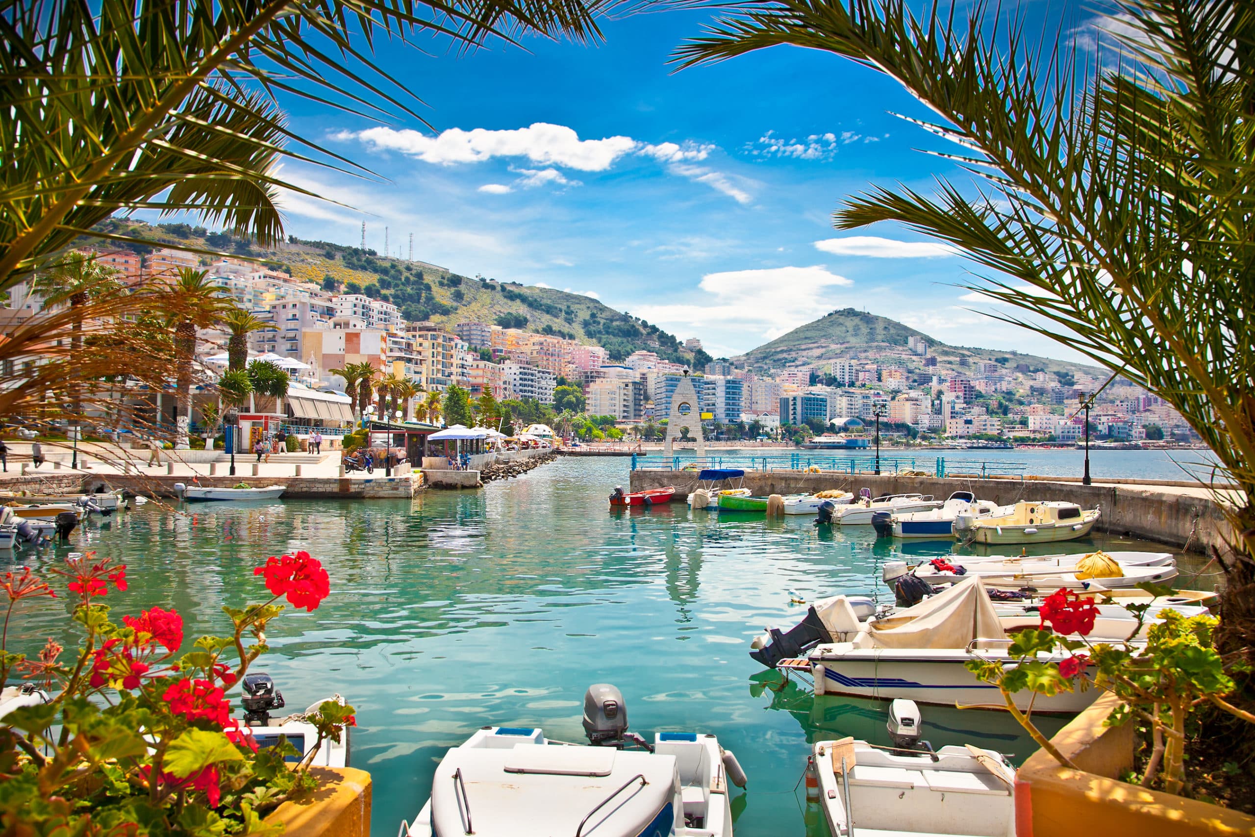  kleine bootjes die in de haven van Sarande in Albanië liggen. Op de voorgrond zie je rode bloemen en palmen en op de achtergrond de boulevard met hoogbouw en daarachter de bergen.