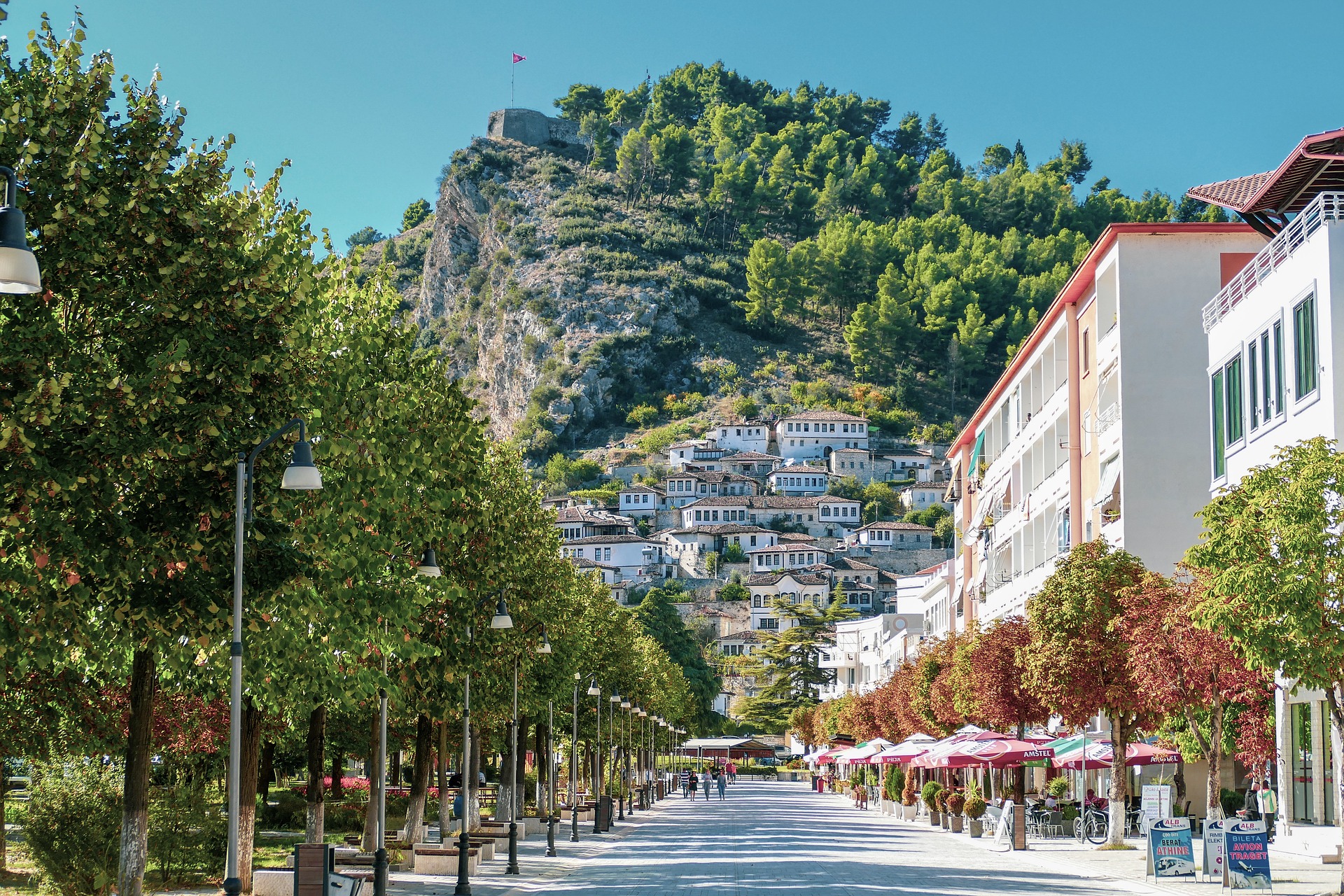 bijna lege straat met aan de linkerkant een bomenrij en aan de rechterkant witte gebouwen en restaurant in de stad Berat in Albanië. Aan het einde van de straat liggen vele witte huisjes tegen een heuvel aan.