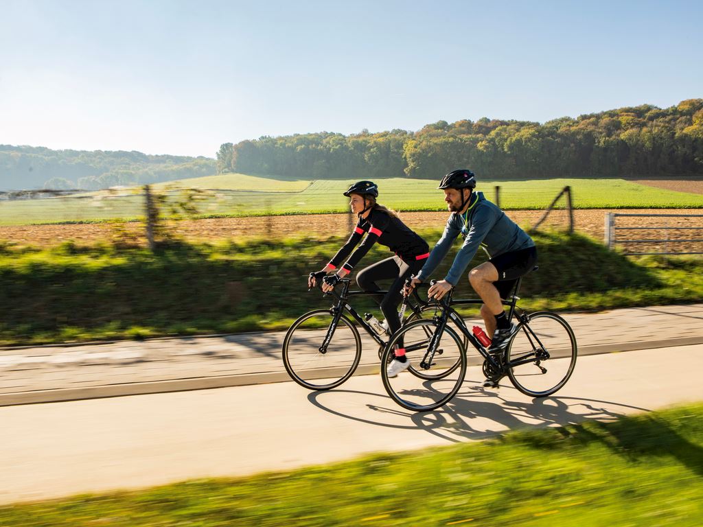 twee wielrenners, een man en een vrouw, die voor velden en een bos langs fietsen in Limburg