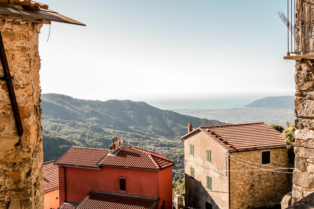 twee huizen in Toscane gezien tussen twee muren door. Eén van de huizen heeft rode muren en de andere heeft muren van steen. Op de achtergrond liggen bergen en platteland en in de verte de zee.