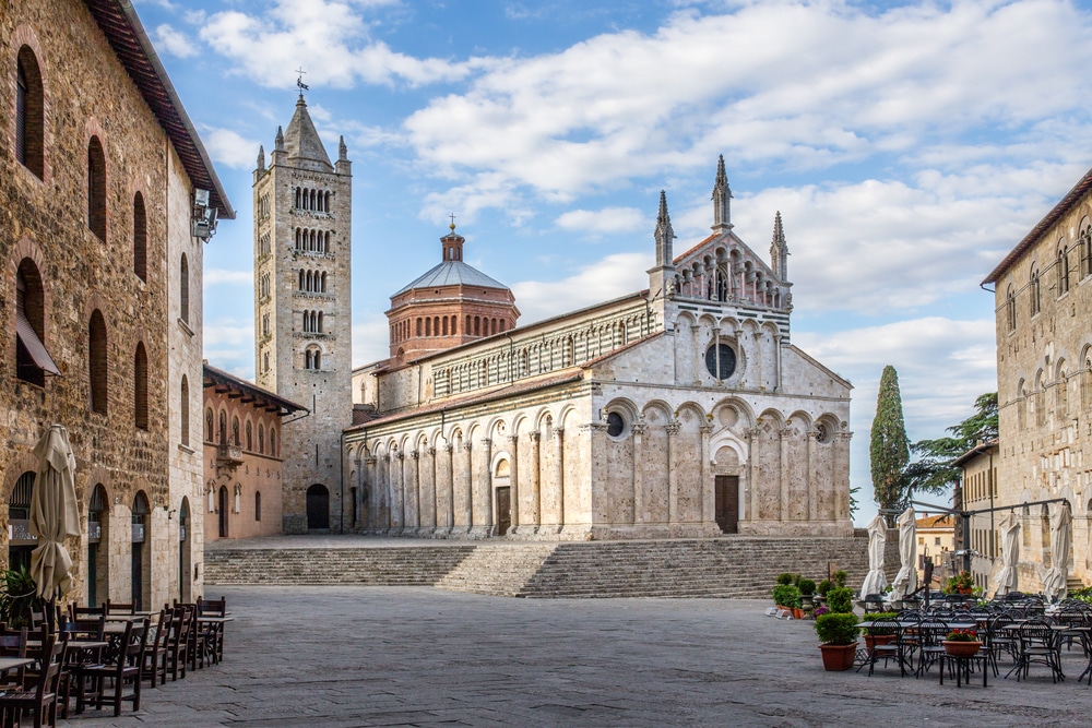 witte kathedraal in het Toscaanse plaatsje Massa Marittima. De kathedraal ligt op een verhoging van een paar treden op een leeg plein.  Aan de zijkanten van het plein staan lege terrassen