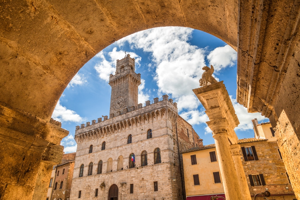 oud, vierkant gebouw met daarbovenop een toren in Montepulciano, gezien van onder een stenen boog vandaan