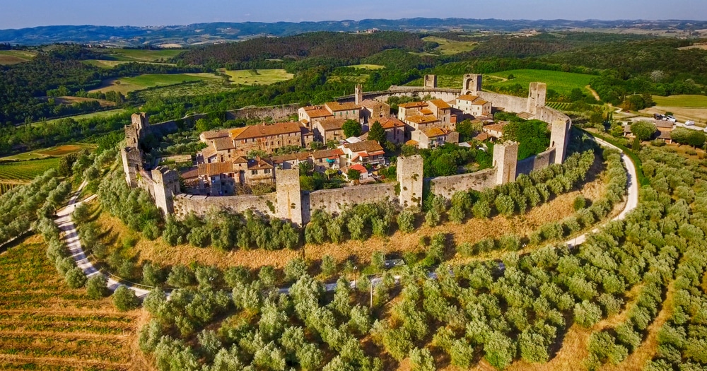 luchtfoto van het ommuurde dorpje Monteriggioni in Toscane. Rondom loopt een pad waar auto's kunnen rijden en liggen bossen en velden.