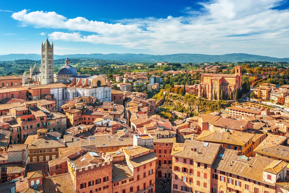 luchtfoto van het oude centrum van Siena met uitzicht over de daken van de huizen en kerken. In de verte zie je de bergen.