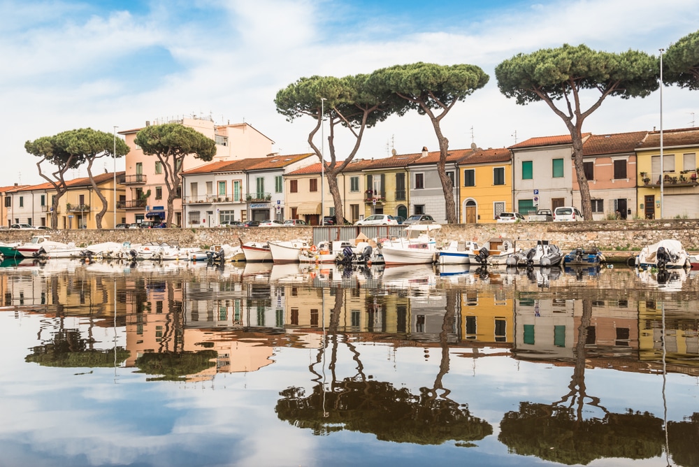 foto van kleine - voornamelijk witte - bootjes in de haven van het Toscaanse plaatsje Viareggio. Daarachter lig een straat met gekleurde huisjes, auto's en een paar bomen. De huizen en bomen weerspiegelen in het water.