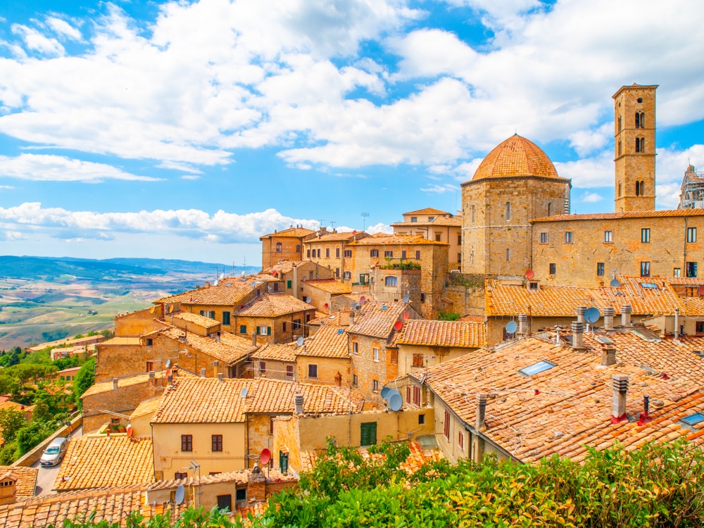het stadje Volterra in de Cecina vallei. Op de foto staan oranje huizen en een toren. Op de achtergrond zie je een weids heuvellandschap.