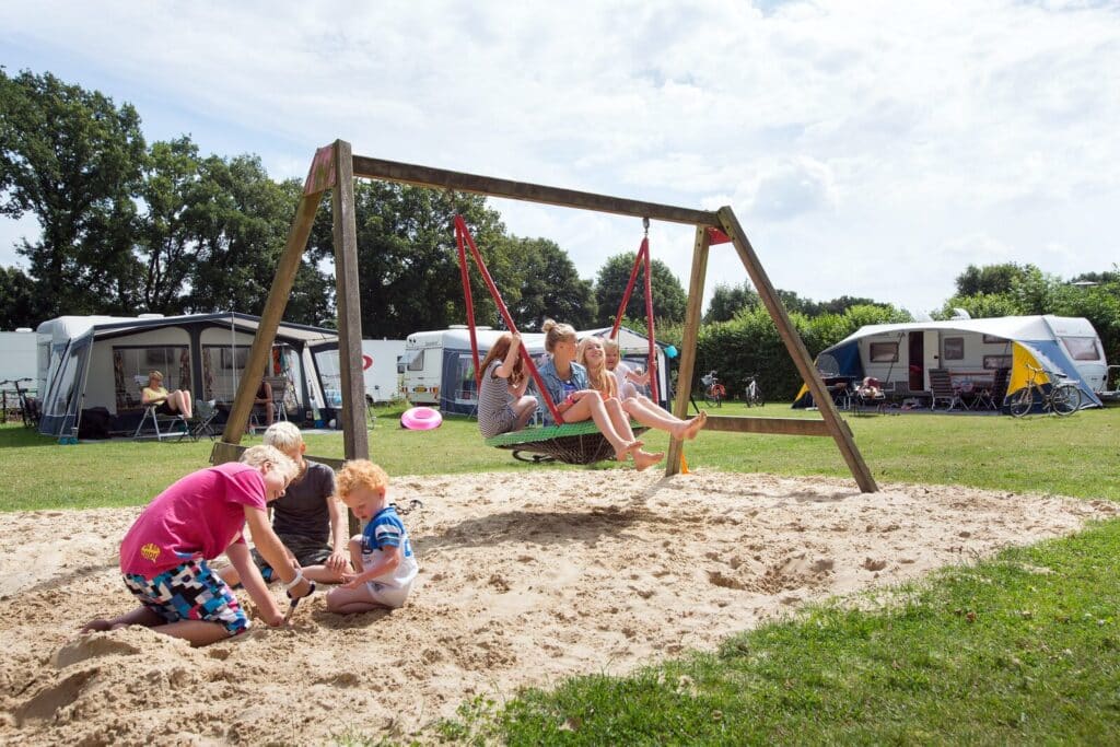 meisjes op een schommel en jongetjes die daarvoor in het zand spelen op Camping De Noetselerberg in Hellendoorn, Overijssel