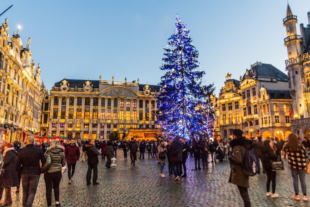 Druk plein in Brussel met grote verlichte kerstboom in het midden. Omringd door gebouwen.