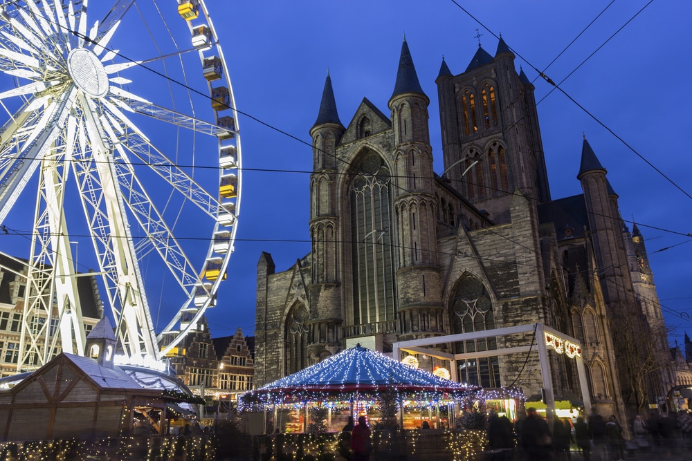 Plein met een grote kerk, verlicht reuzenrad en versierde kraampjes in Gent.