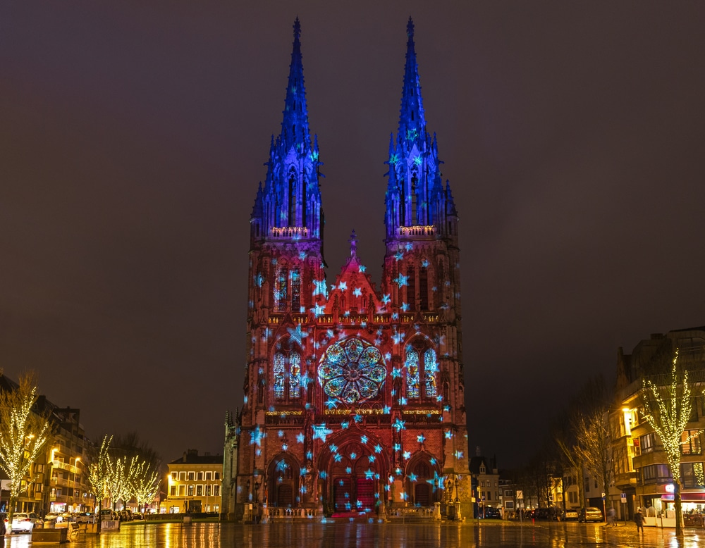 Grote met kerstlichtjes versierde kerk op een plein in Oostende. Omringd door gebouwen en verlichte bomen.