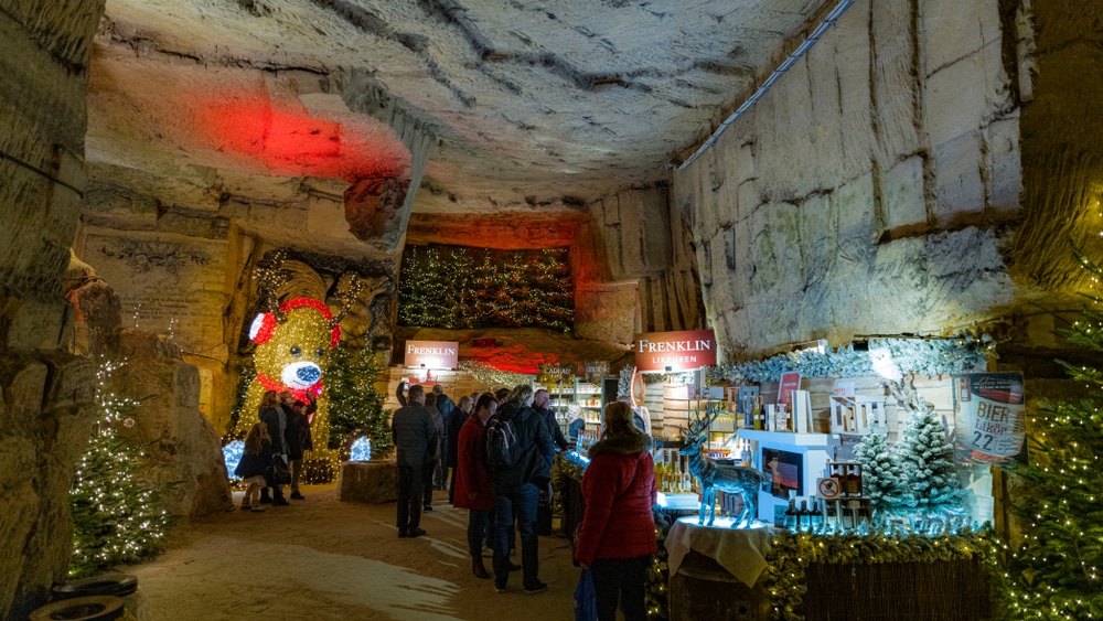 mensen in winterkleding die kijken naar kraampjes tijdens een kerstmarkt in een grot in Valkenburg. De grot is versierd met kerstlichtjes, kerstbomen en een grote verlicht beer met koptelefoon