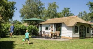 man met een zoontje die op het grasveld naast een rieten cottage voetballen. Daarnaast staat een moeder en zit een dochtertje aan de tafel op het terras.