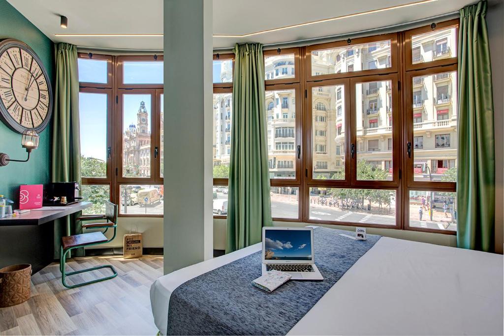 laptop op het bed van een hotelkamer met grote ramen die uitzicht bieden op het Plaza del Ayutnamiento in Valencia